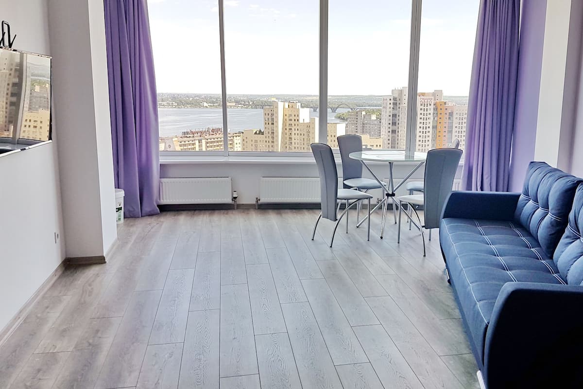 A modern living room with a blue sofa, a dining table with chairs, and a view of a cityscape and river.