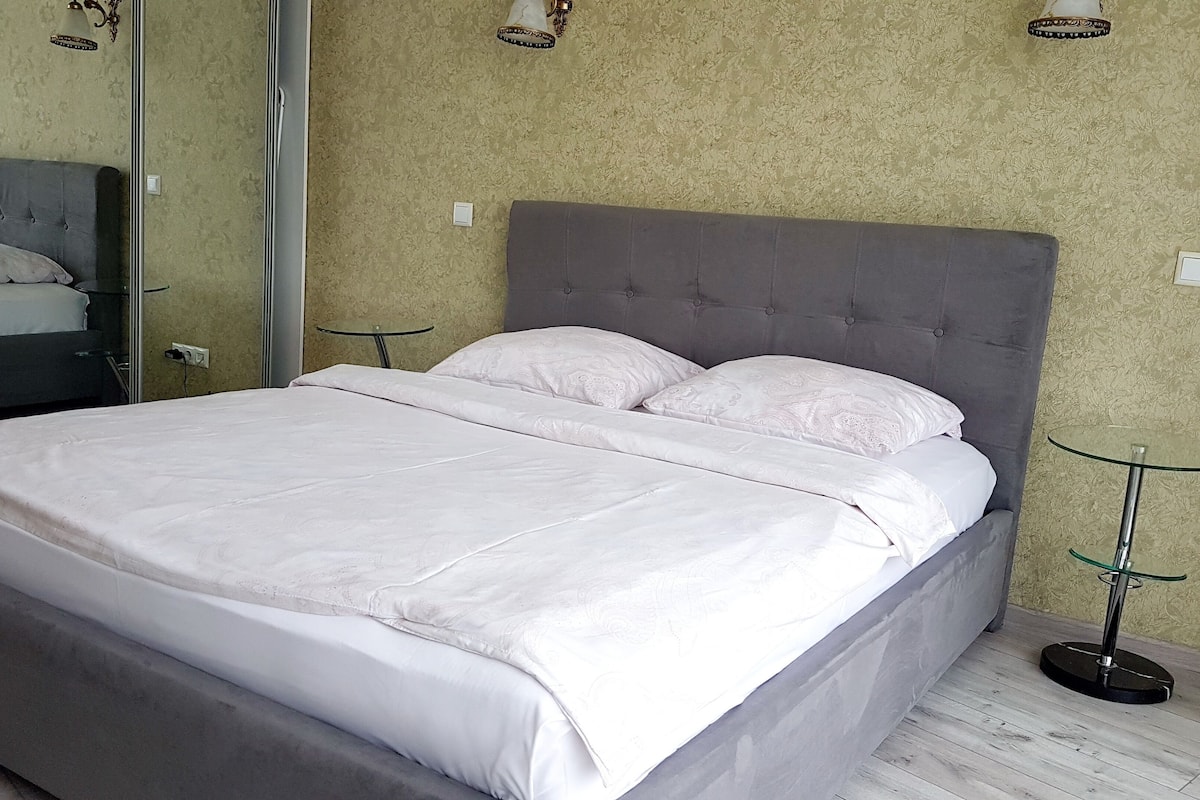 A neatly made bed with a grey headboard, a small glass table beside it, and a mirror on the wall.
