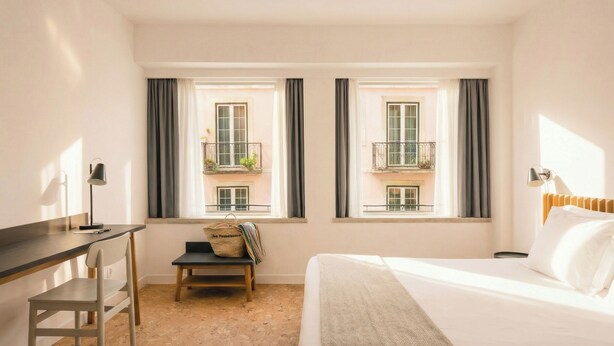 A hotel room with a bed, desk, chair, and two windows with balconies.