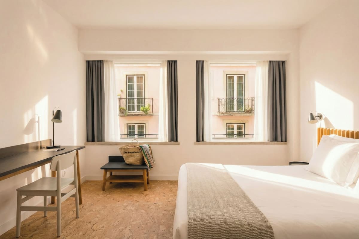 Een hotelkamer met een bed, bureau, stoel en twee ramen met balkons.