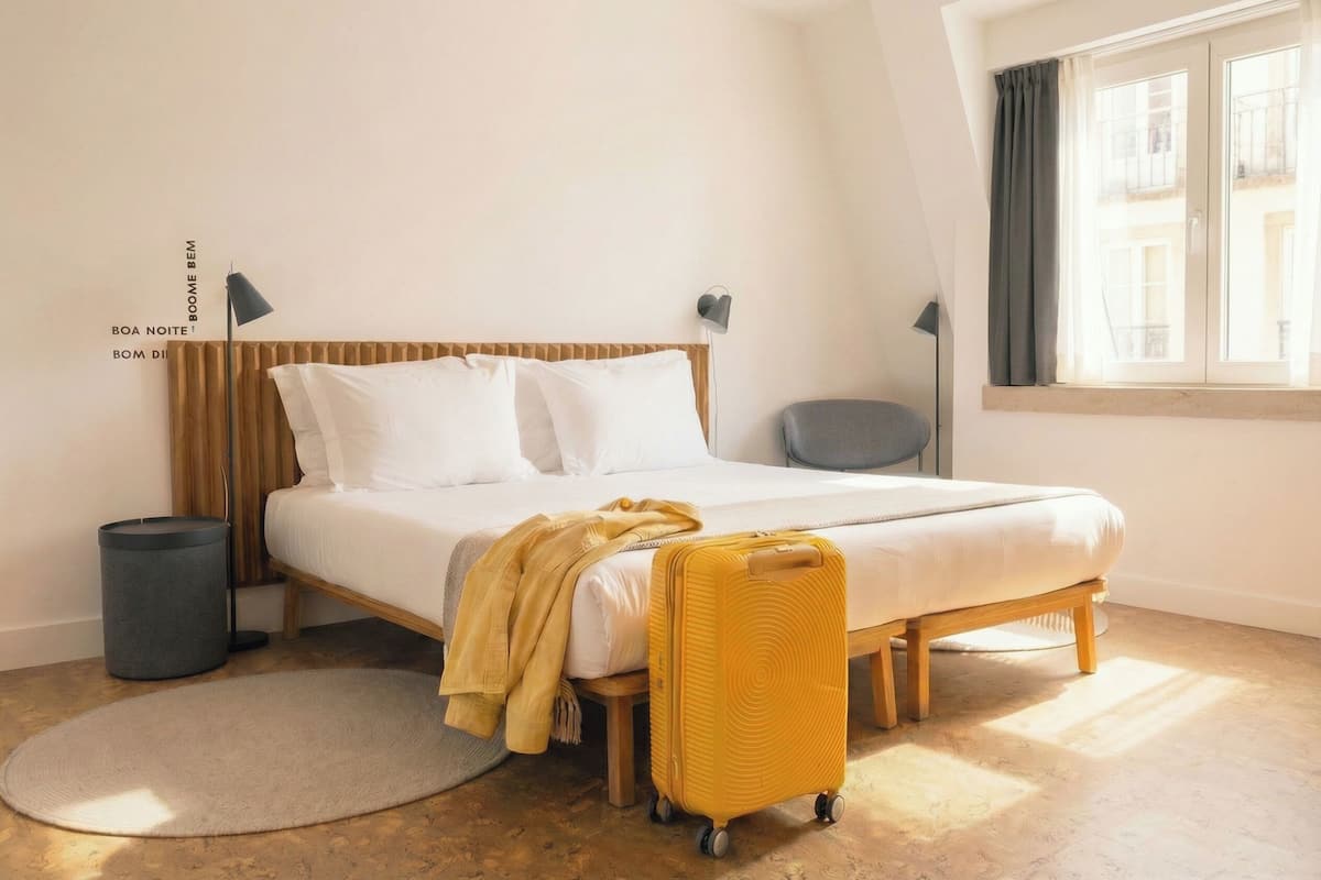 Een net opgemaakt bed met witte lakens en een gele deken, een houten hoofdbord, een klein nachtkastje met een lamp, een rond vloerkleed, en een koffer op de vloer.