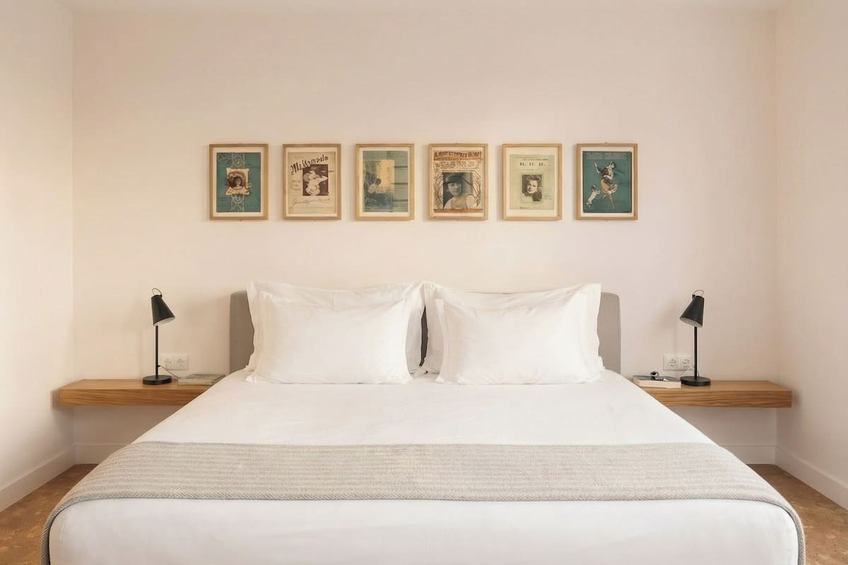 Een opgemaakt bed met witte lakens, twee nachtkastjes met lampen, en vijf ingelijste foto's aan de muur.
