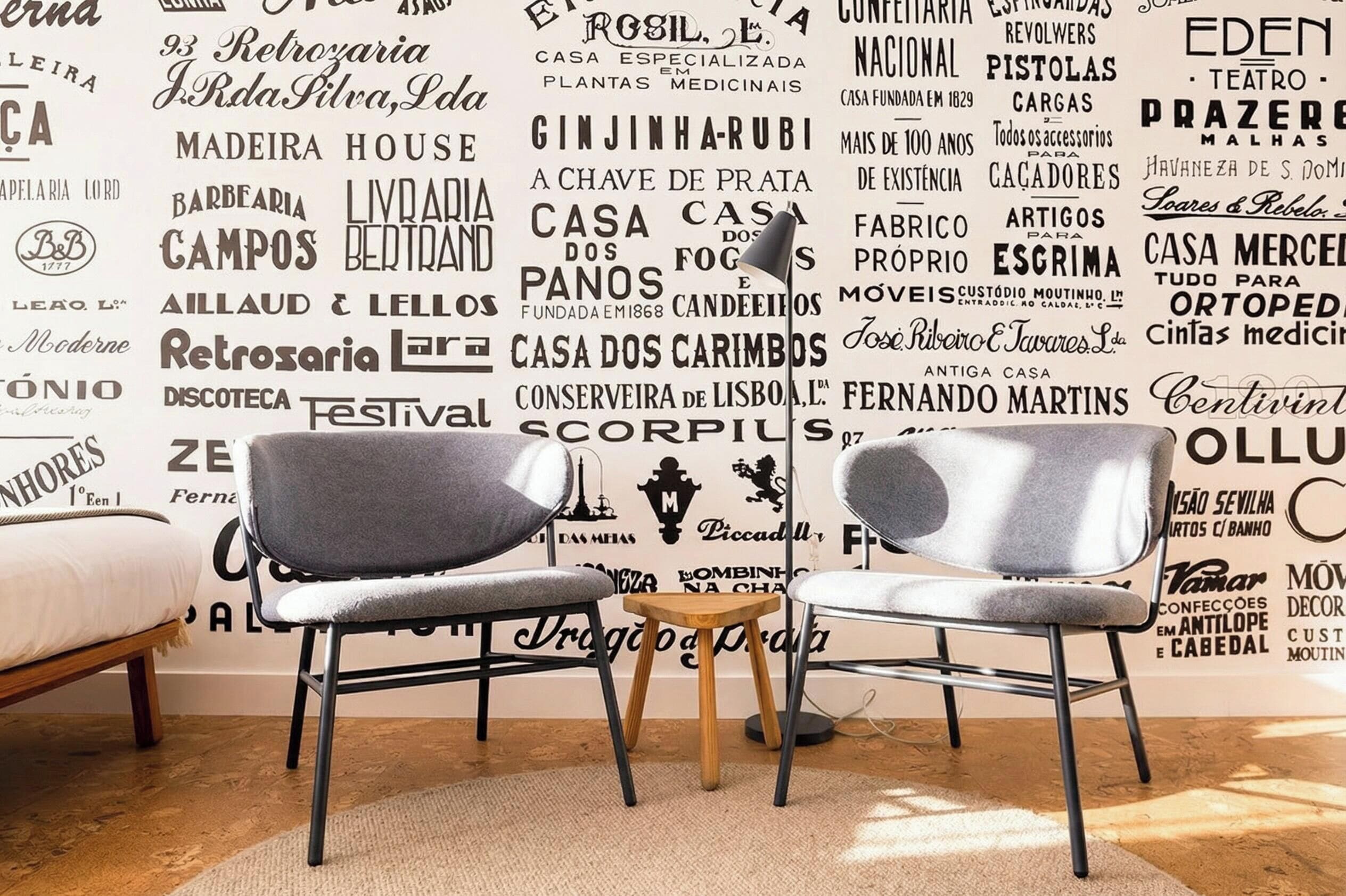 Una habitación con dos sillas grises, una mesita de madera y una pared decorada con varias palabras y frases portuguesas.