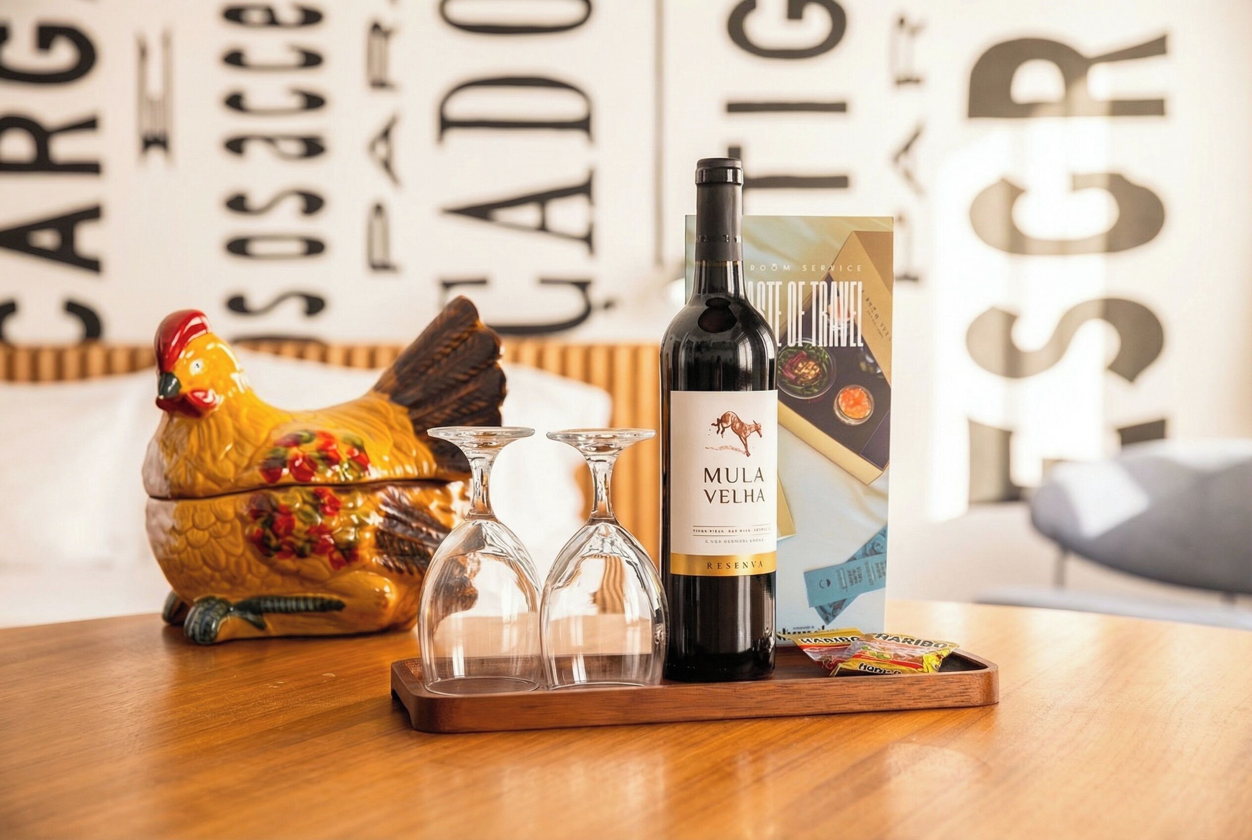 Una bottiglia di vino Mula VELHA, due bicchieri da vino, un gallo in ceramica decorativa e una scatola di caramelle gommose Haribo su un tavolo di legno.