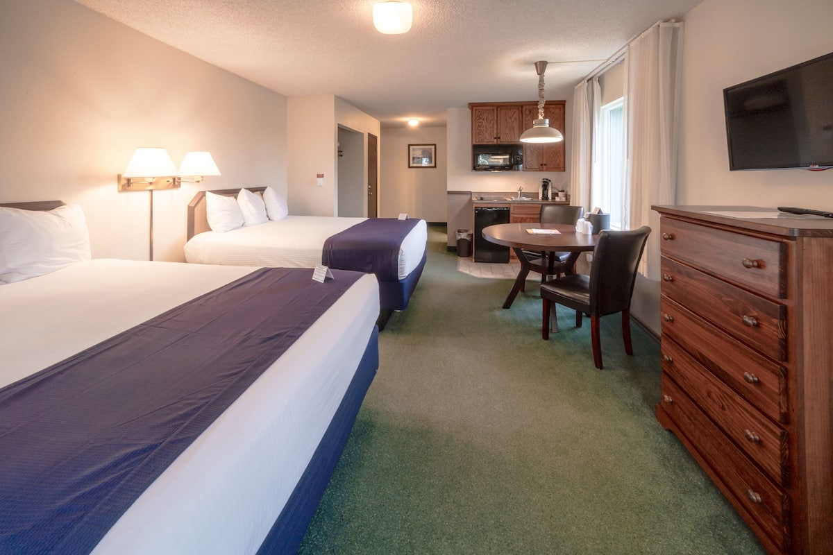 A hotel room with two beds, a desk, a chair, a television, and a kitchenette.