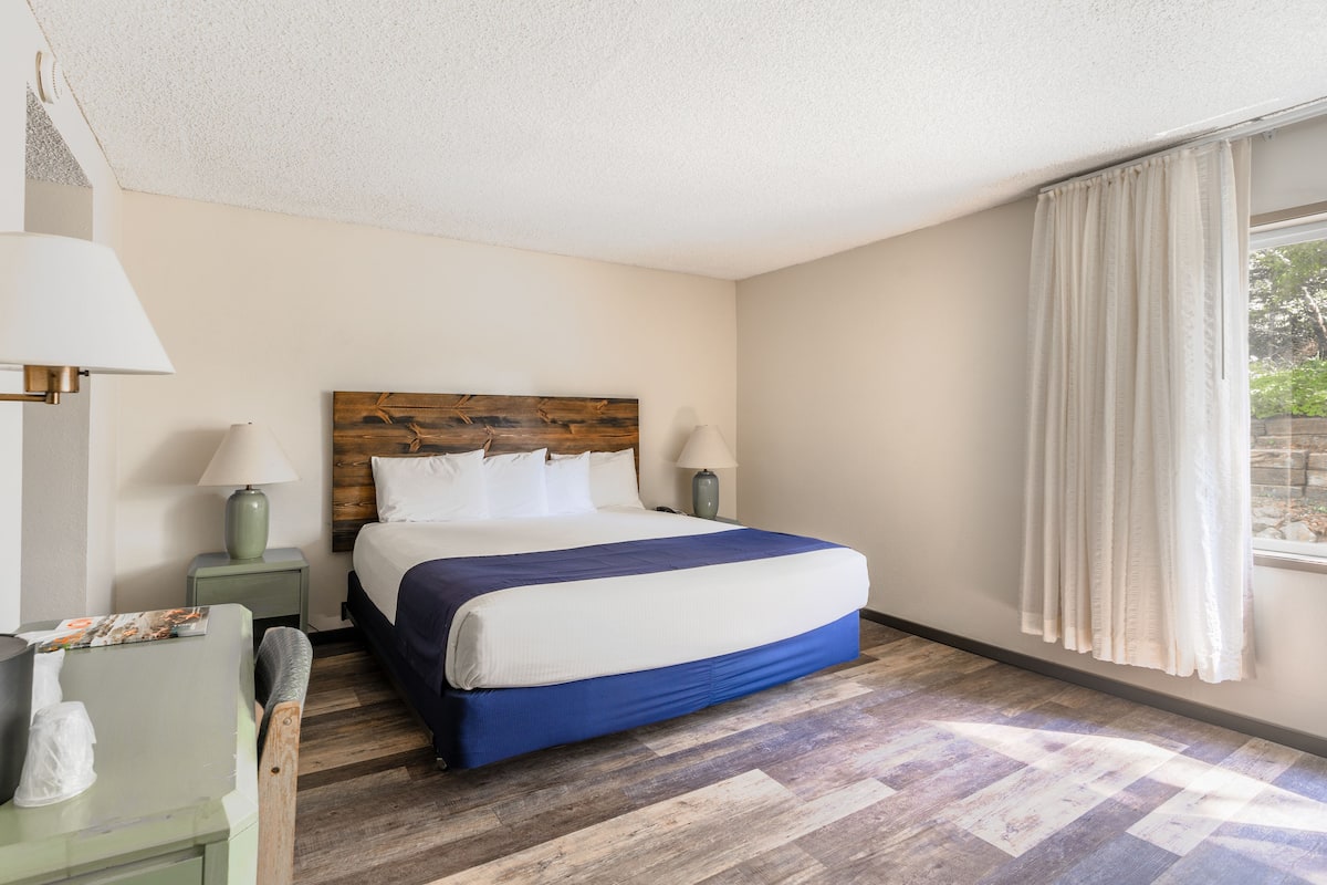A bedroom with a wooden headboard, a bed with white and blue bedding, two bedside lamps, a nightstand, and a window with curtains.