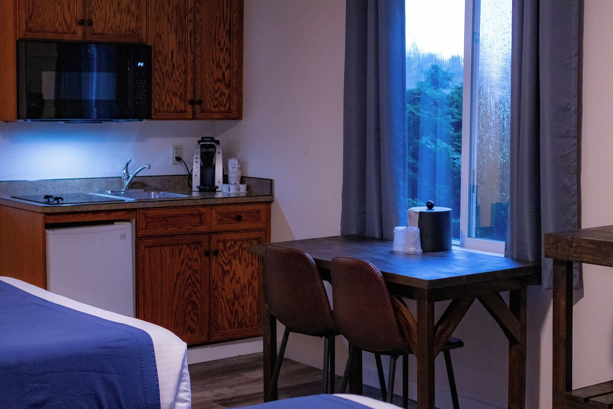 A compact hotel room with a kitchenette, a bed with a blue blanket, a wooden table with chairs, and a view through a window.