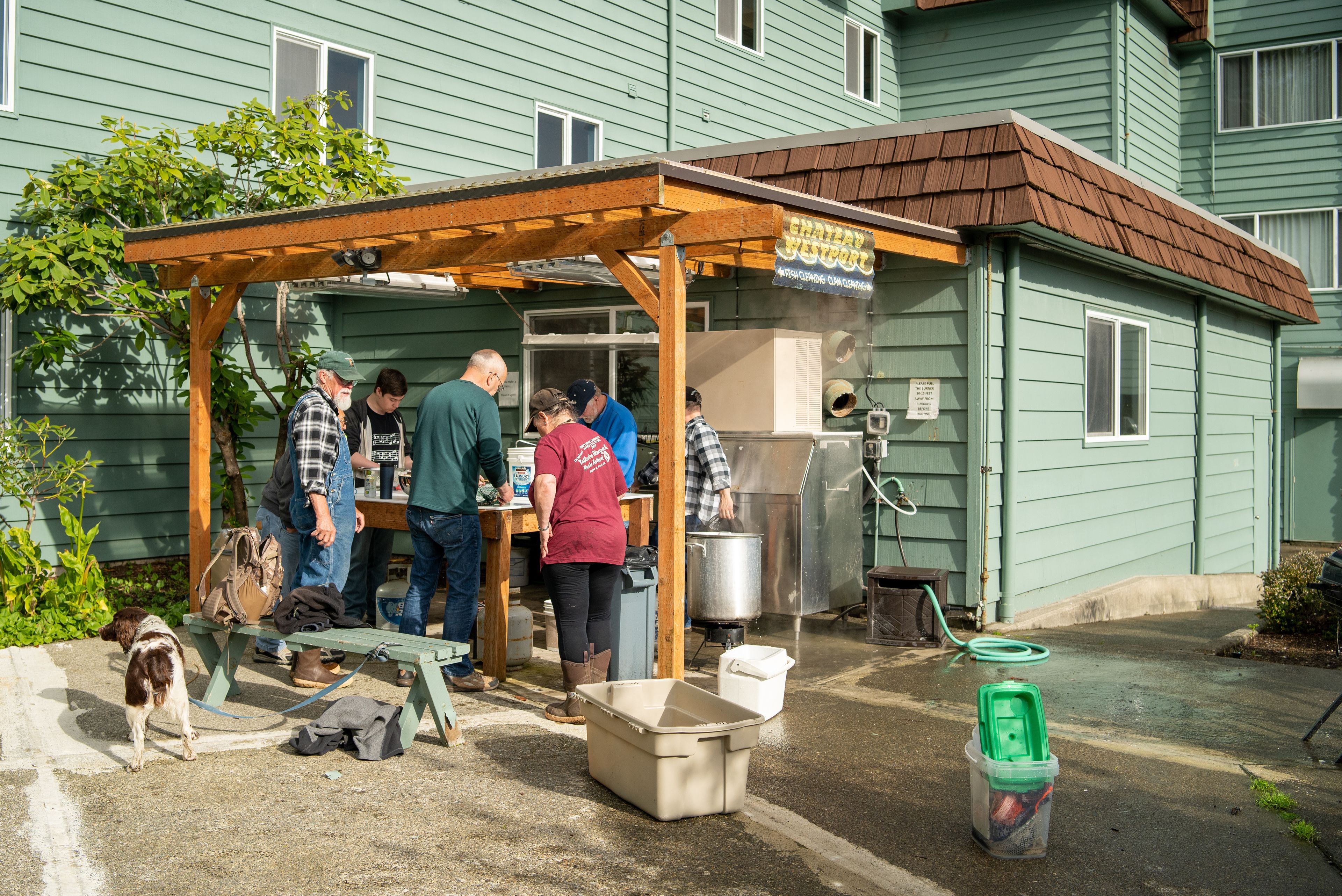 A group of people gathered around a wooden structure with a sign, outside a green building with a brown roof.