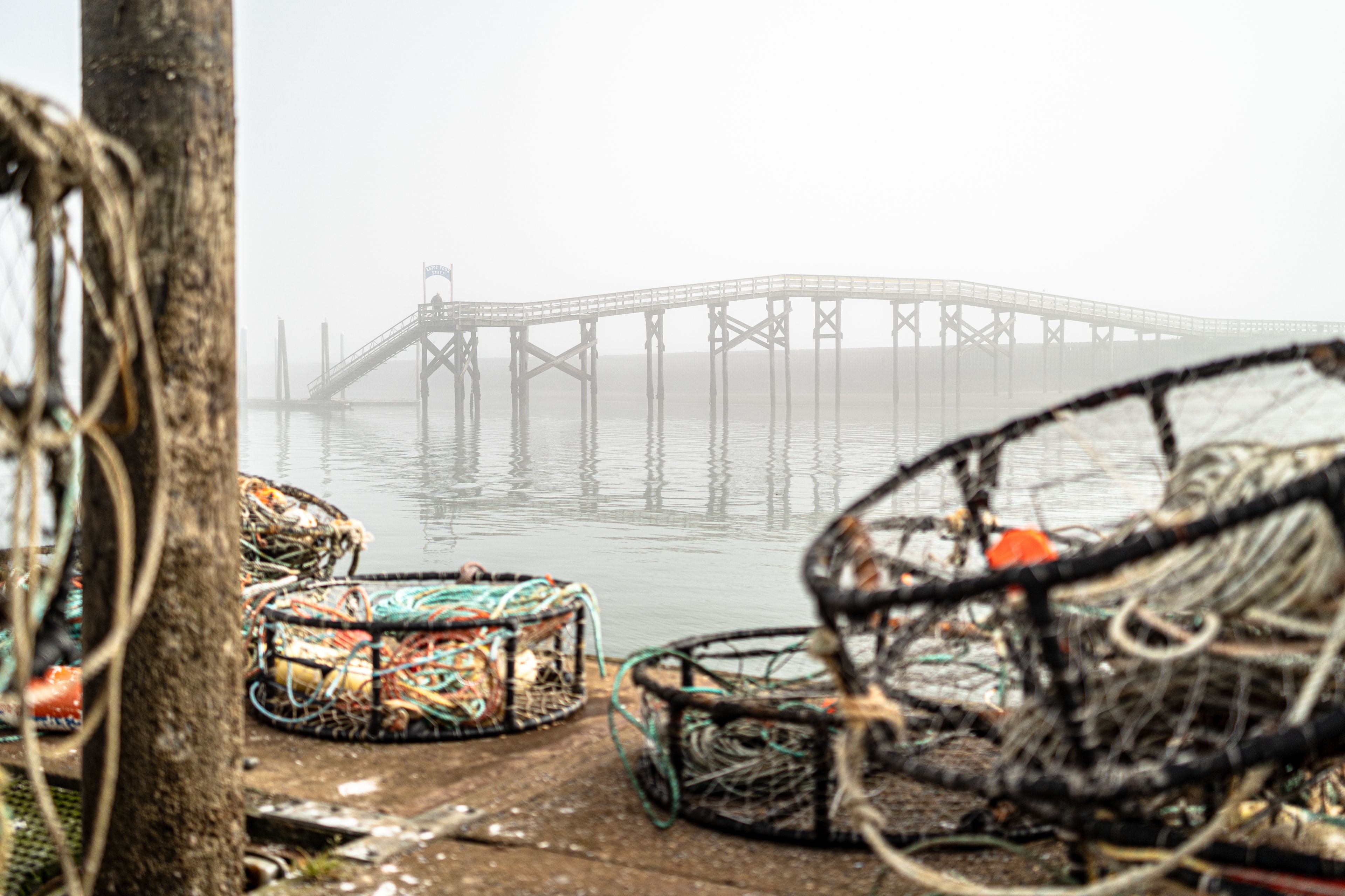 Escena costera brumosa con equipos de pesca y un muelle al fondo.