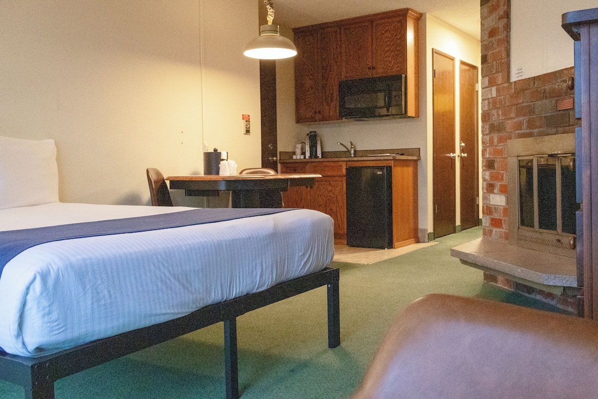 A hotel room with a bed, a desk, a chair, a kitchenette, and a fireplace.