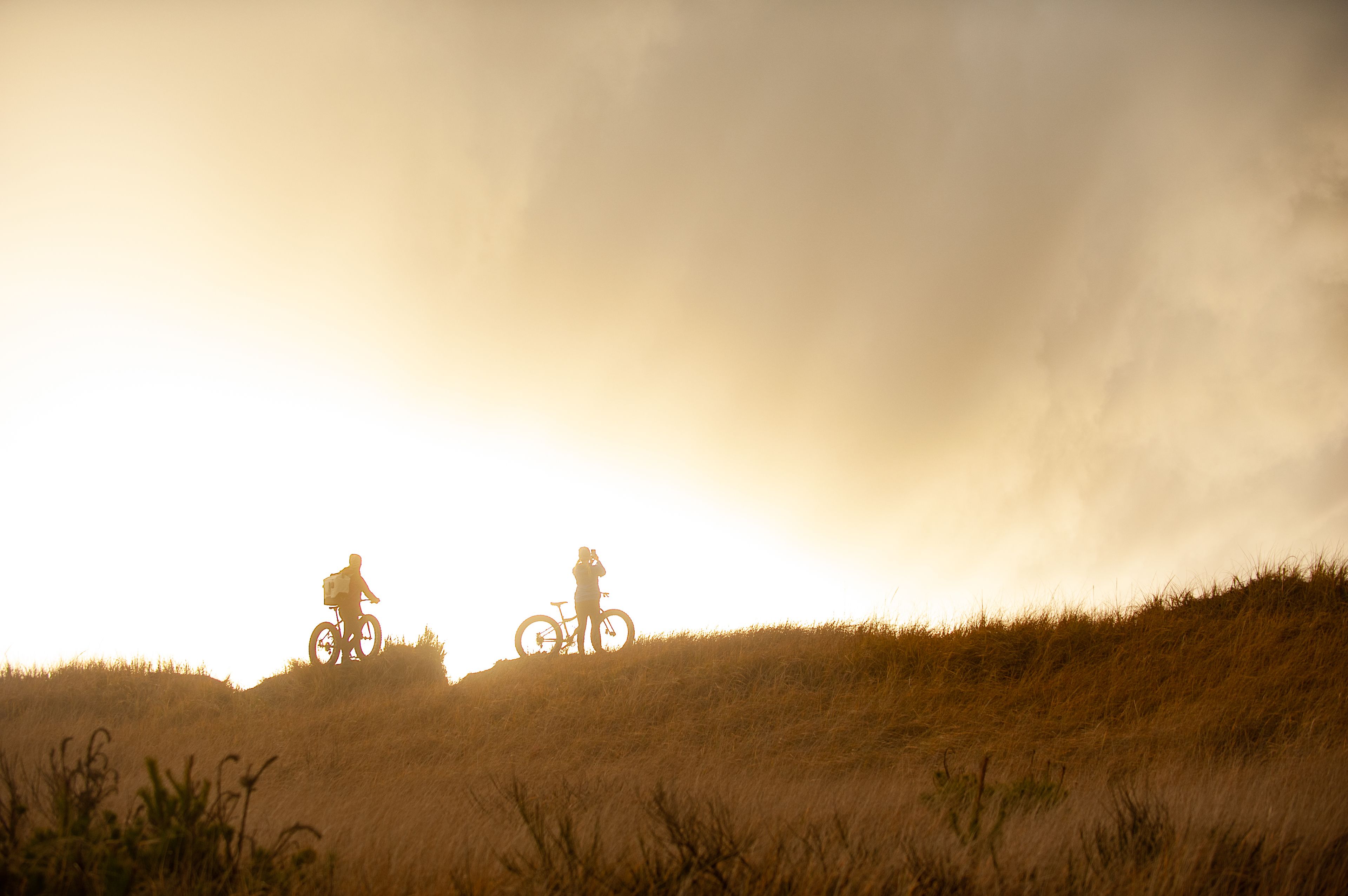 A silhouette of a person on a bicycle on a grassy hill with a cloudy sky.