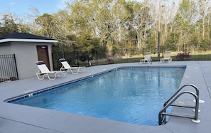 A rectangular swimming pool with a tiled edge, surrounded by a low black metal fence. There are two lounge chairs and a small building with a door and window to the left.