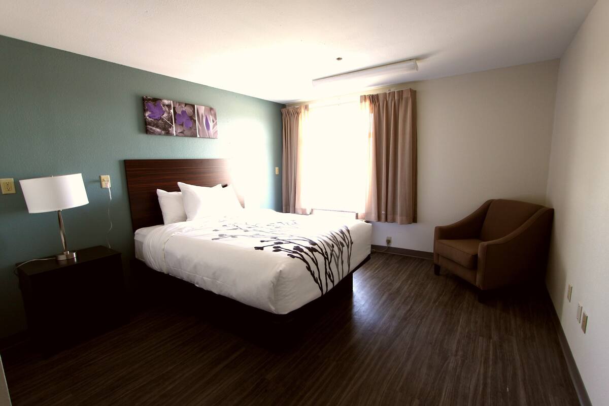 Una habitación de hotel con cama, una silla, una mesita de noche, una lámpara y una ventana con cortinas.