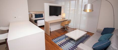 Una habitación de hotel moderna con suelo de madera, un sofá, un escritorio con televisor, una silla y una lámpara.