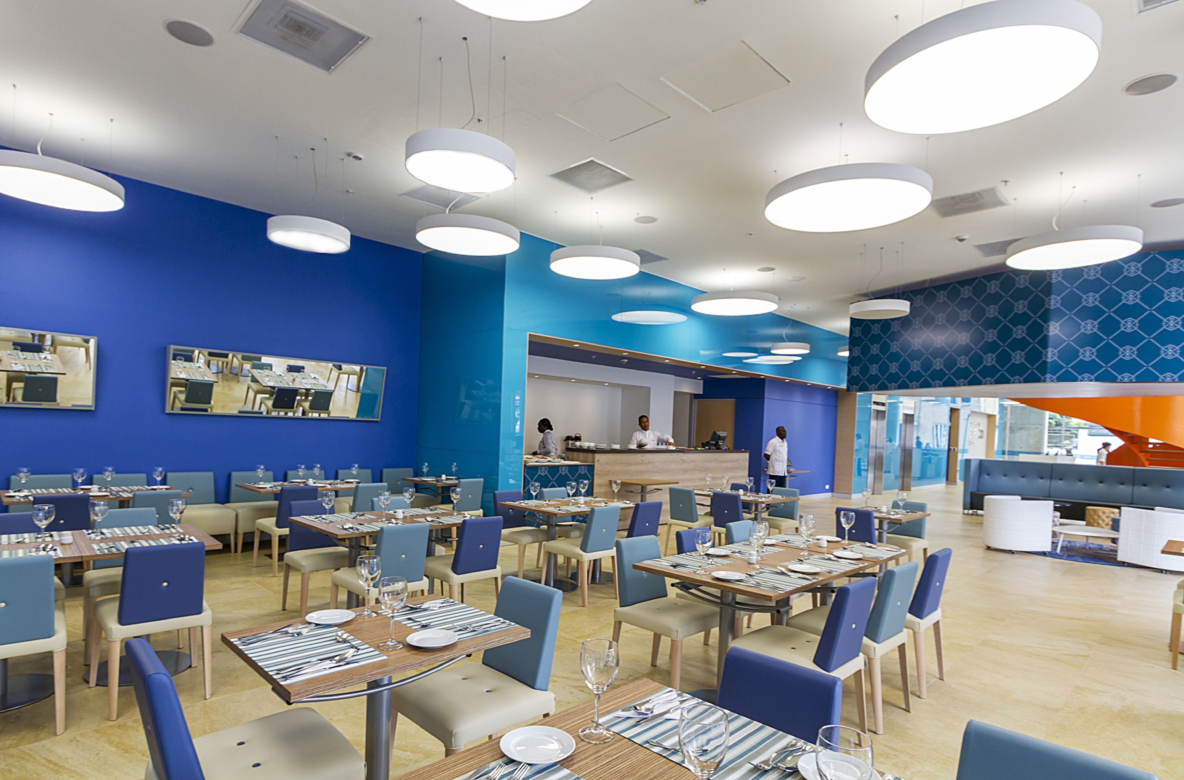 Ein modernes Restaurant mit blauen Wänden und mehreren Essbereichen, mit runden Deckenleuchten und einer zentralen Theke.