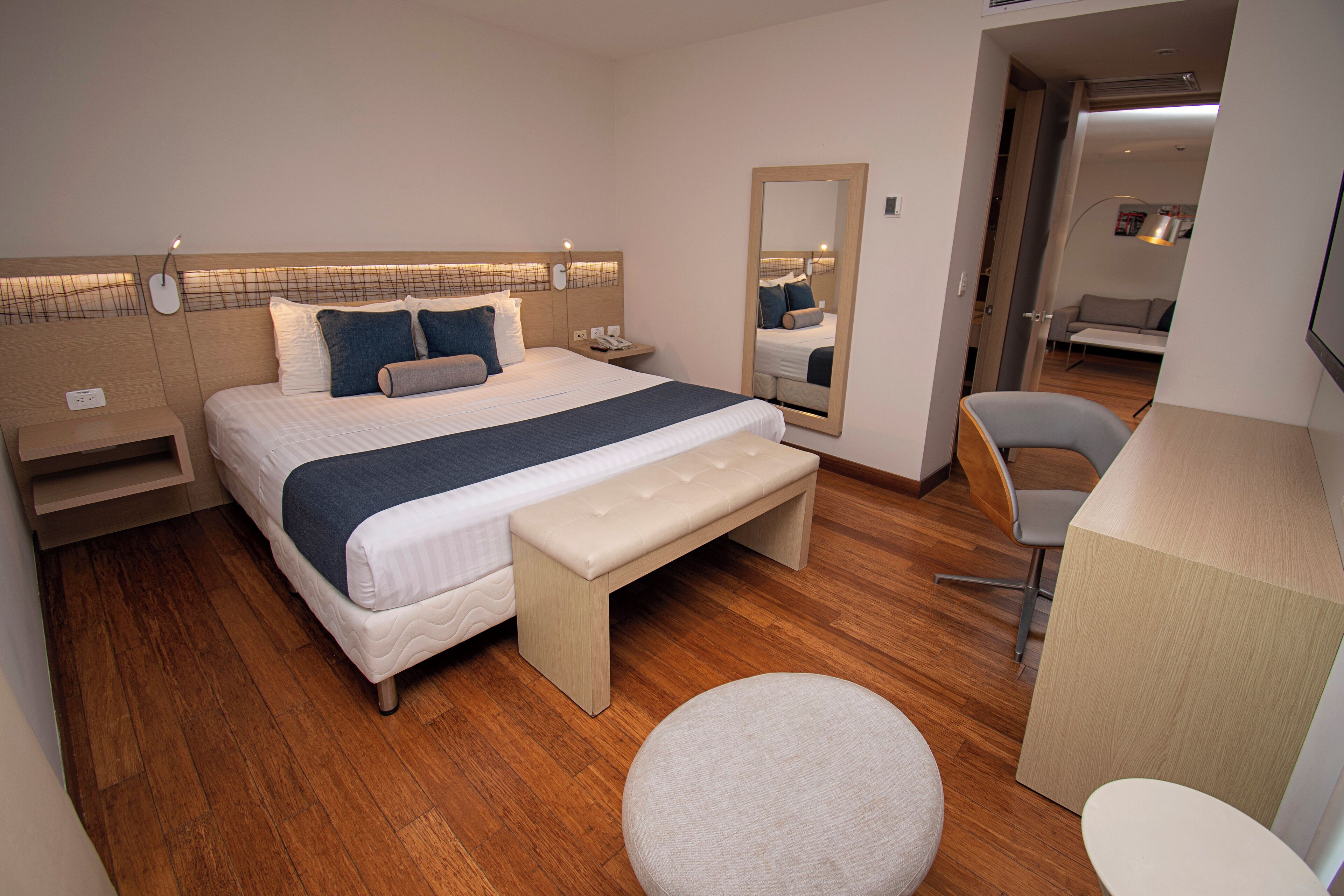 A modern hotel room with a large bed, bedside tables, a bench, a round ottoman, a chair, and a desk.