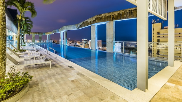 An indoor swimming pool with a view of the city at night, surrounded by palm trees and modern architecture. - Cosmos Pacifico Hotel (Buenaventura)