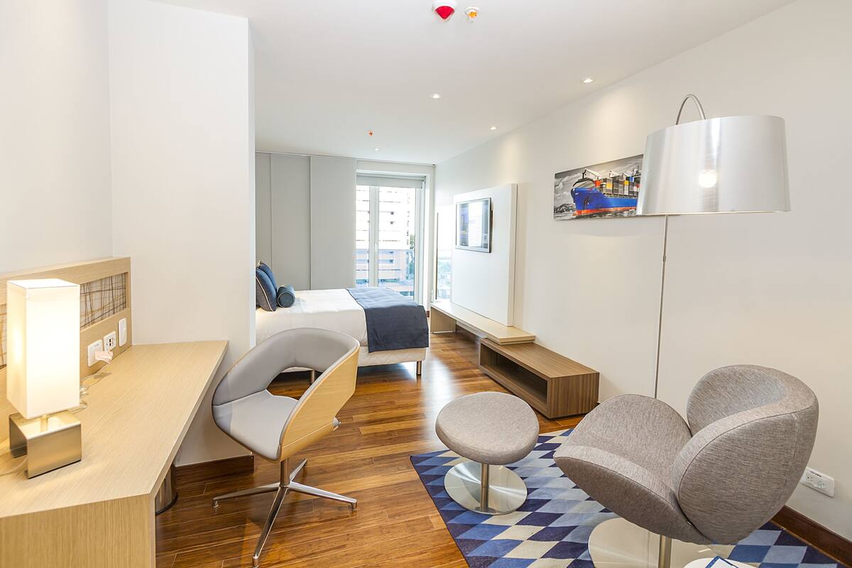 Een moderne hotelkamer met een houten bureau, een comfortabele stoel, een bed met een blauwe hoofdbord, een bankje en een groot raam met uitzicht op de stad.
