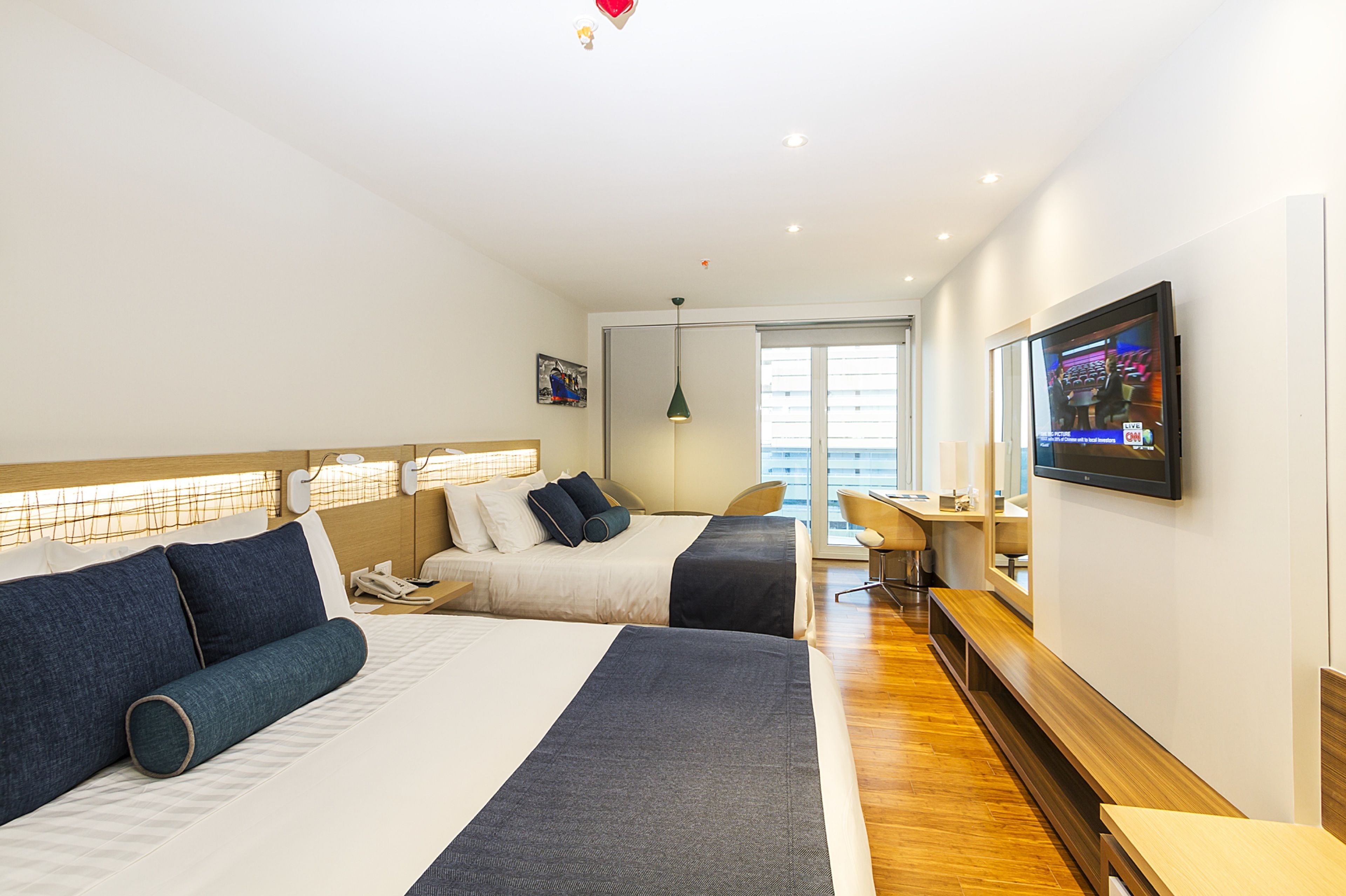 A hotel room with two beds, a desk, a TV, and a window with blinds.