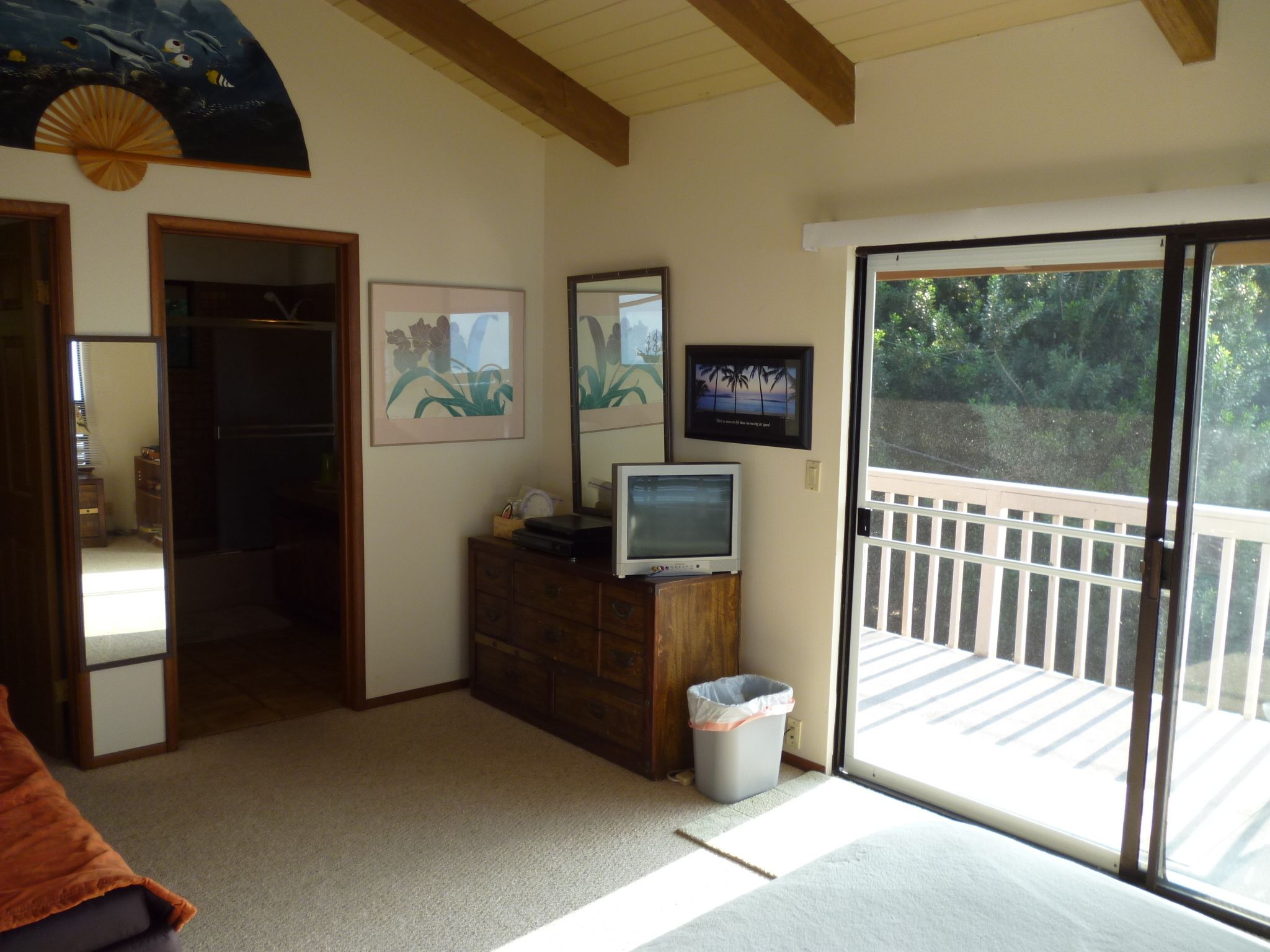 A room with a sliding glass door leading to a balcony, a wooden dresser, a television, and framed artwork on the wall.