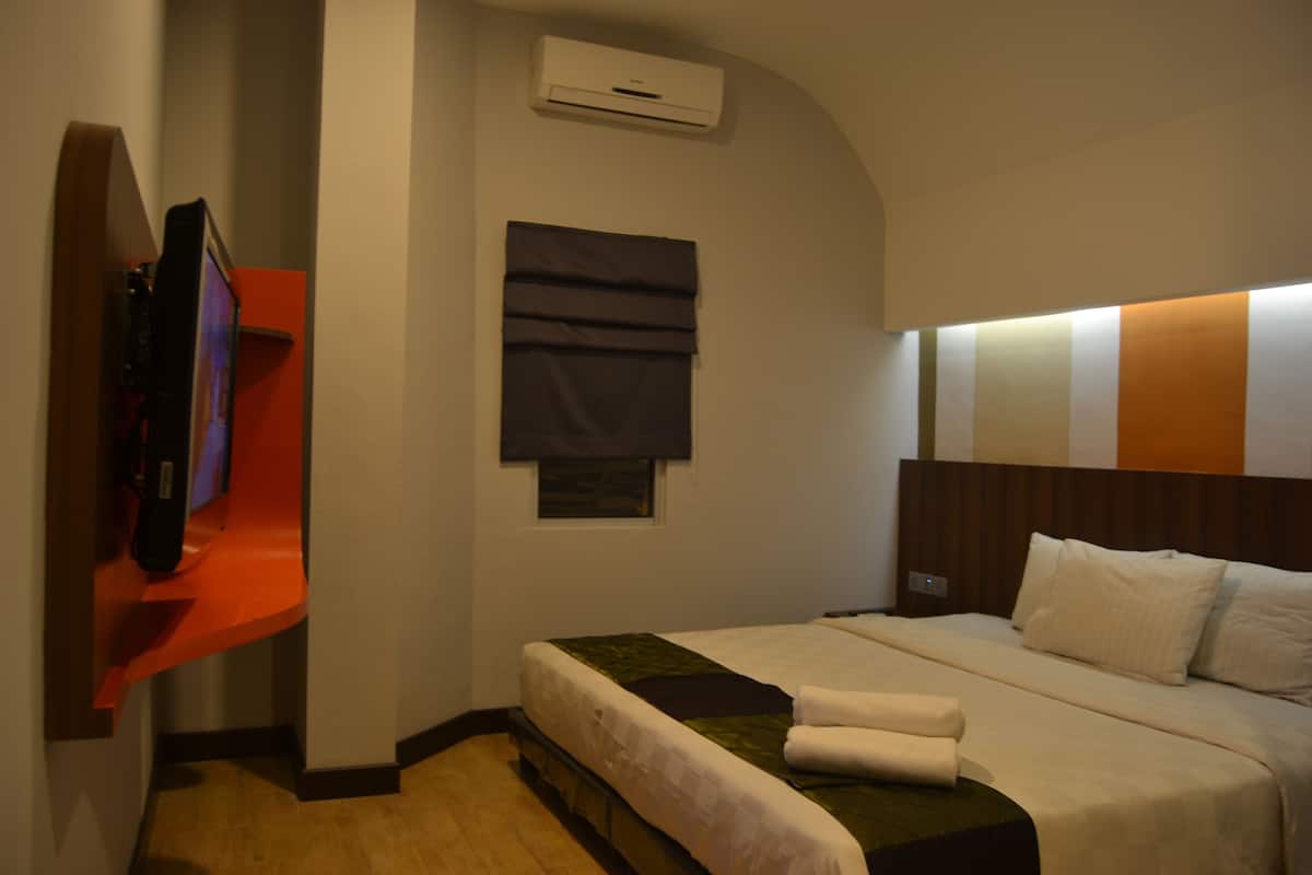 A hotel room with a bed, a flat-screen TV mounted on a red wall, a window with dark curtains, and a wall-mounted air conditioner.