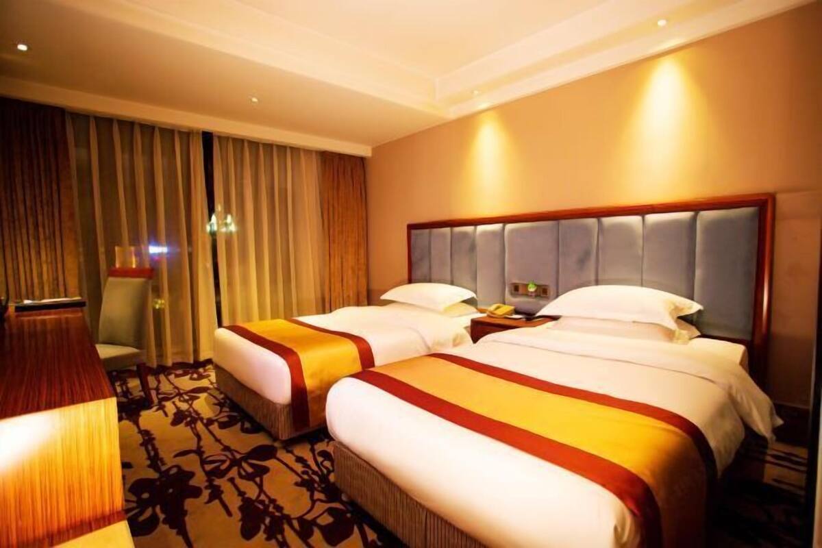 A hotel room with two beds, a large window with curtains, a wooden headboard, and a carpeted floor.