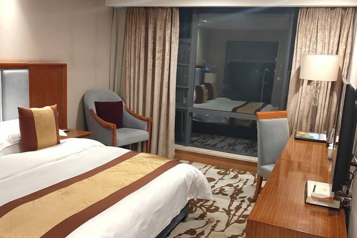 A hotel room with a large bed, a desk, two chairs, a TV, and a large window with curtains.