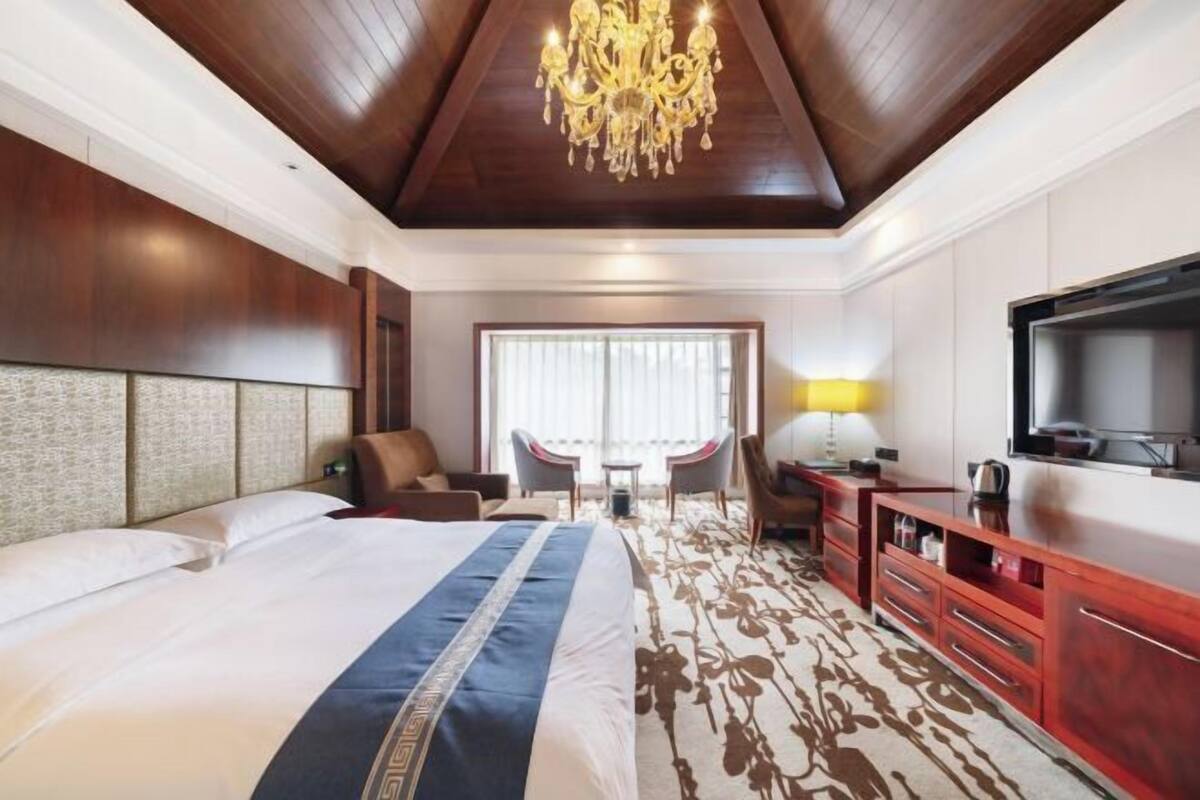 A hotel room with a large bed, a chandelier, a TV, and a desk with a lamp.