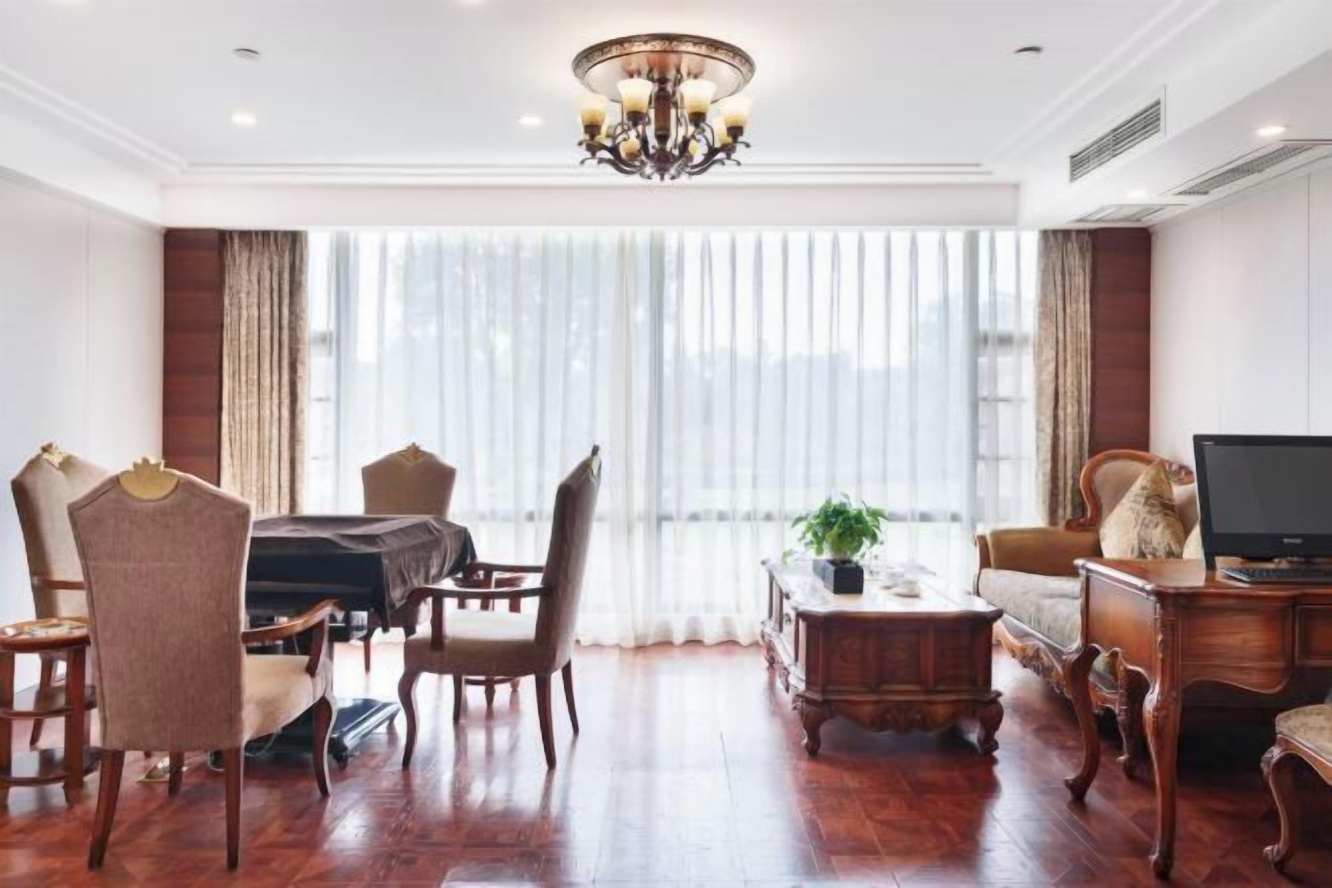 A spacious living room with a large window, a chandelier, wooden furniture, and a television.