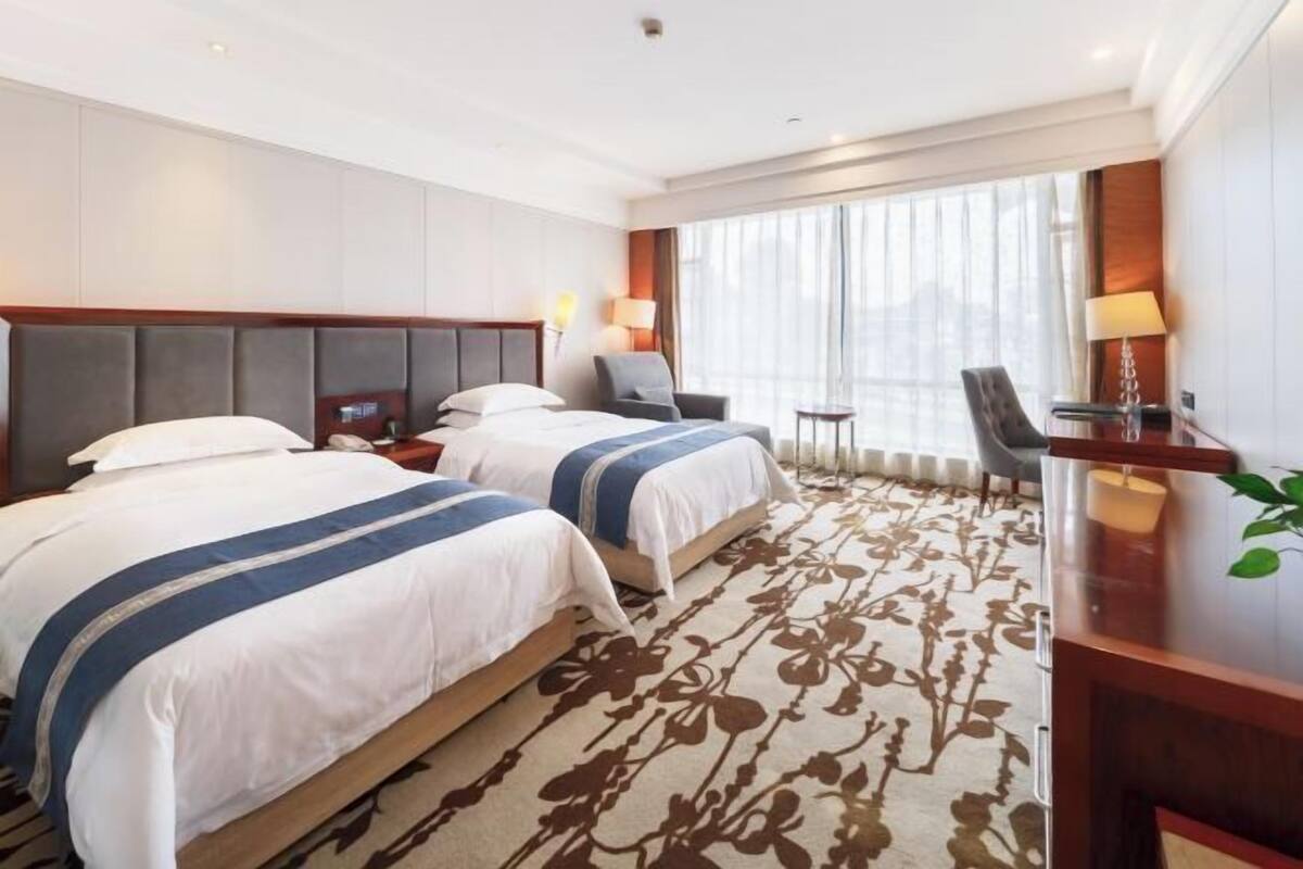 A hotel room with two beds, a desk, a chair, a lamp, and a large window with curtains.