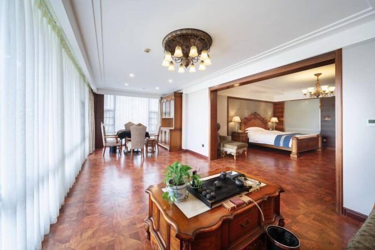 A hotel room with a wooden floor, a dining area with chairs, a bed with a blue blanket, and a chandelier.