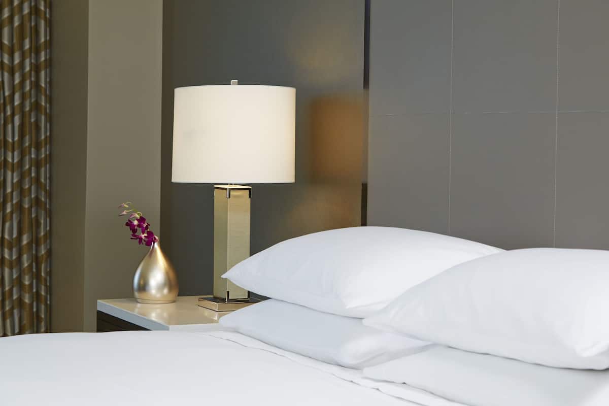 Una cama bien tendida con sábanas y almohadas blancas, una mesita de noche con un florero y una lámpara con pantalla blanca.