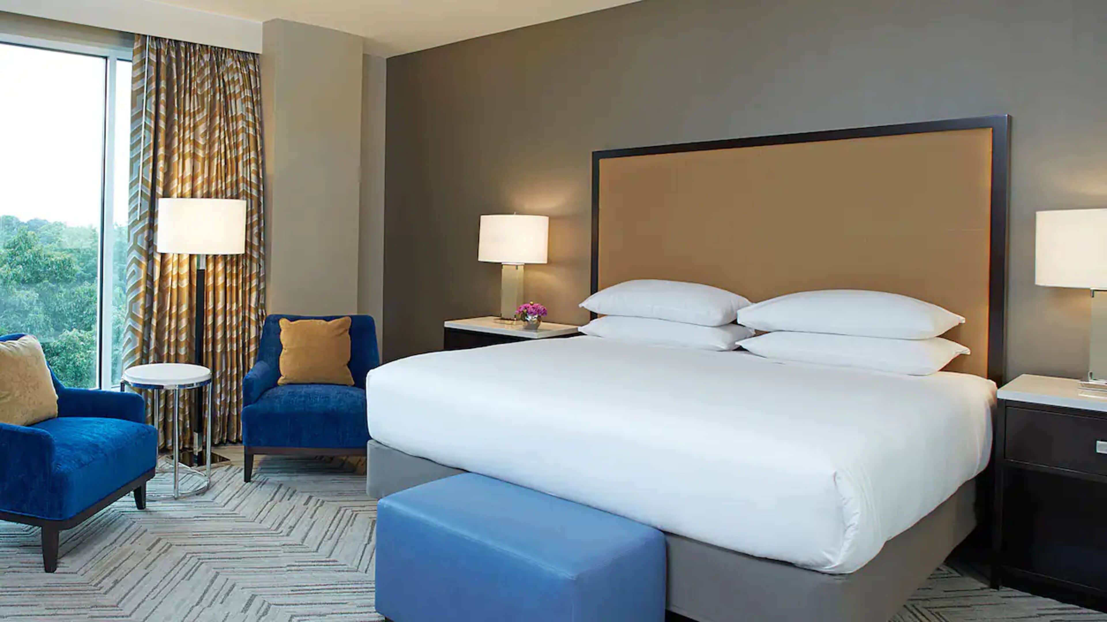 A hotel room with a large bed, two bedside tables with lamps, a blue armchair, a small round table, and a view of trees through the window.