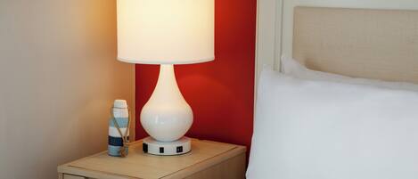 Una cama bien hecha con sábanas blancas, una lámpara de noche y una botella de agua a rayas sobre una mesita de madera.