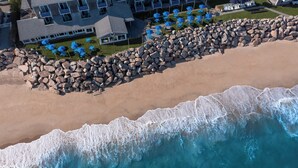 An aerial view of a beach with a rocky shoreline, a building with multiple balconies, and blue umbrellas. - Pleasant View Inn (Westerly)
