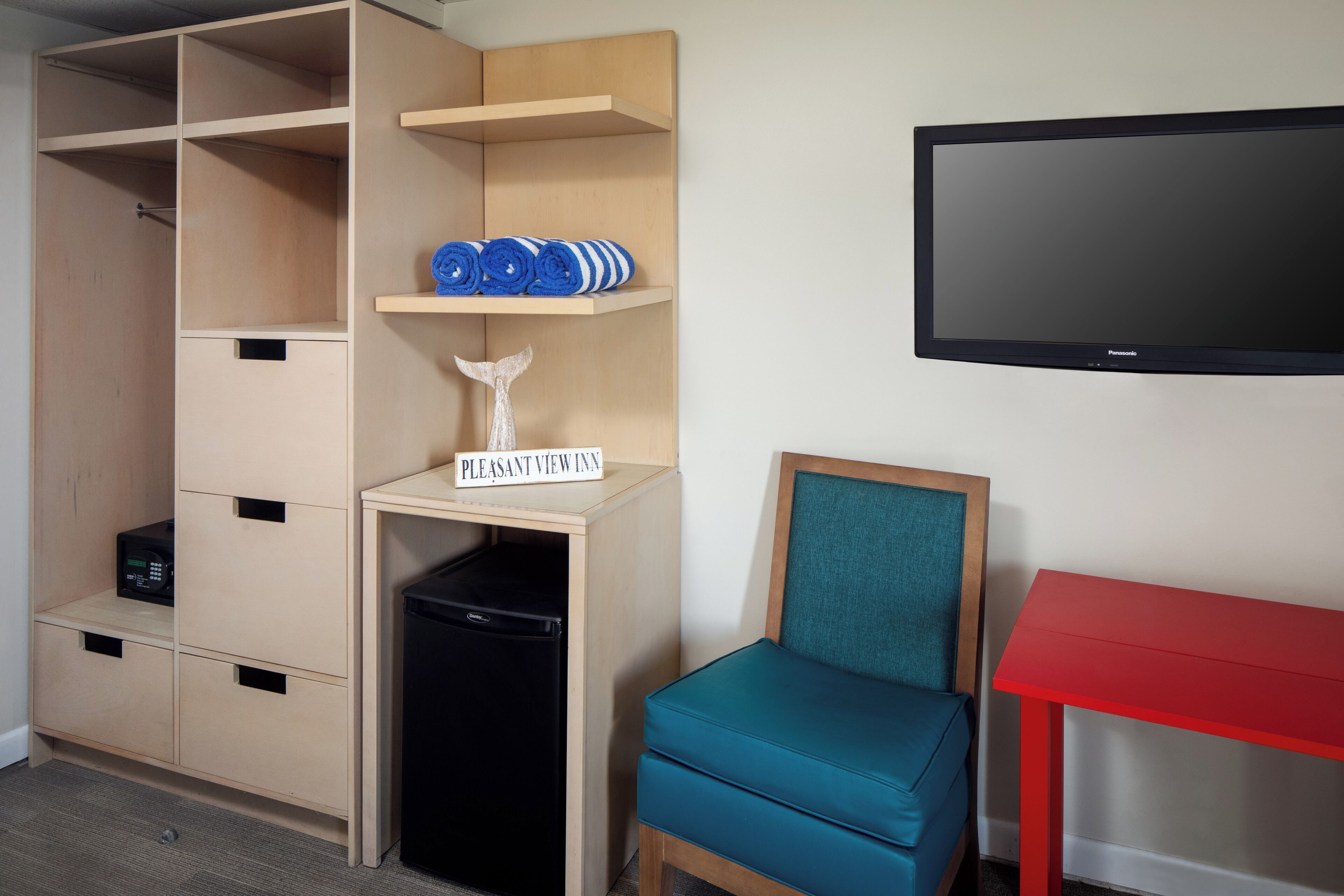 A room with a wooden wardrobe, a small black appliance, a teal chair, a red table, and a mounted TV.