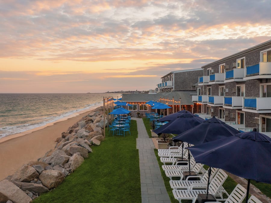 A beachfront hotel with outdoor seating and umbrellas, a stone wall, and a view of the ocean.