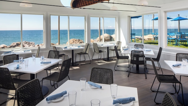 An outdoor dining area with tables and chairs, large windows overlooking the sea, and a wooden ceiling structure. - Pleasant View Inn (Westerly)