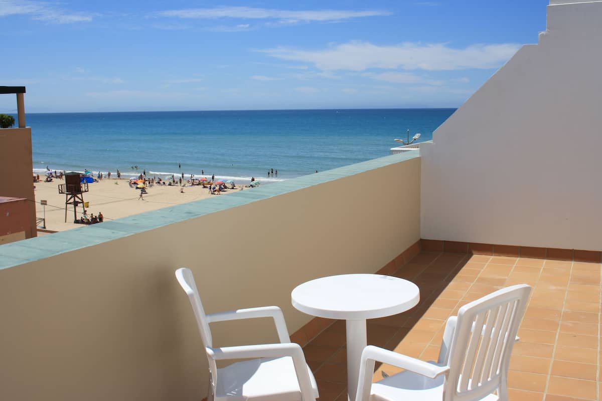 A balcony with a view of the beach and ocean. There are two white chairs and a small round table.
