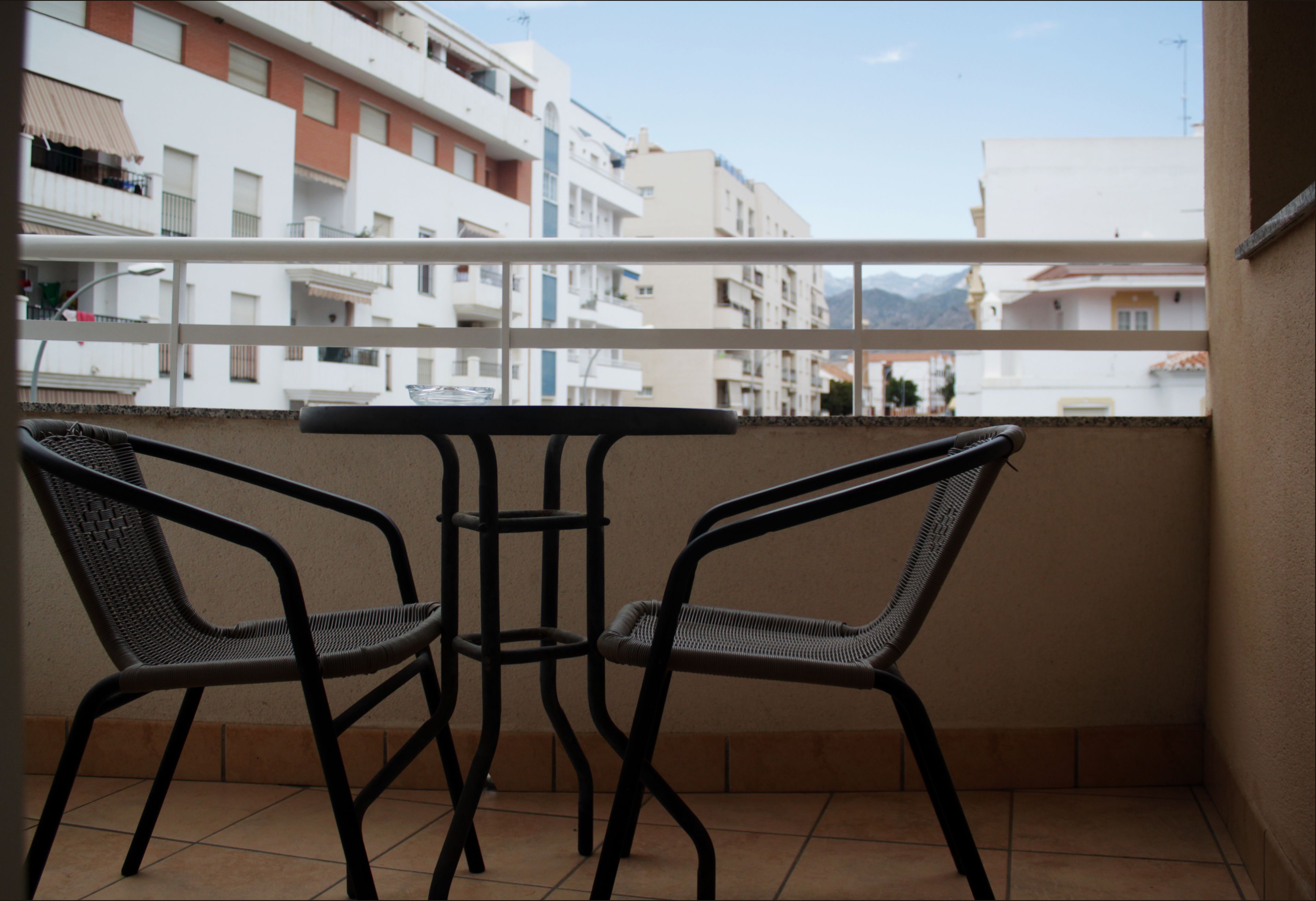 A balcony with two wicker chairs and a small table, overlooking a residential area with white buildings and a mountain in the distance.