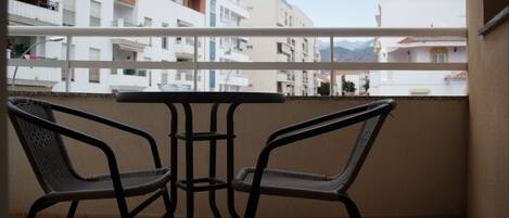 A balcony with two wicker chairs and a small table, overlooking a residential area with white buildings and a mountain in the distance.