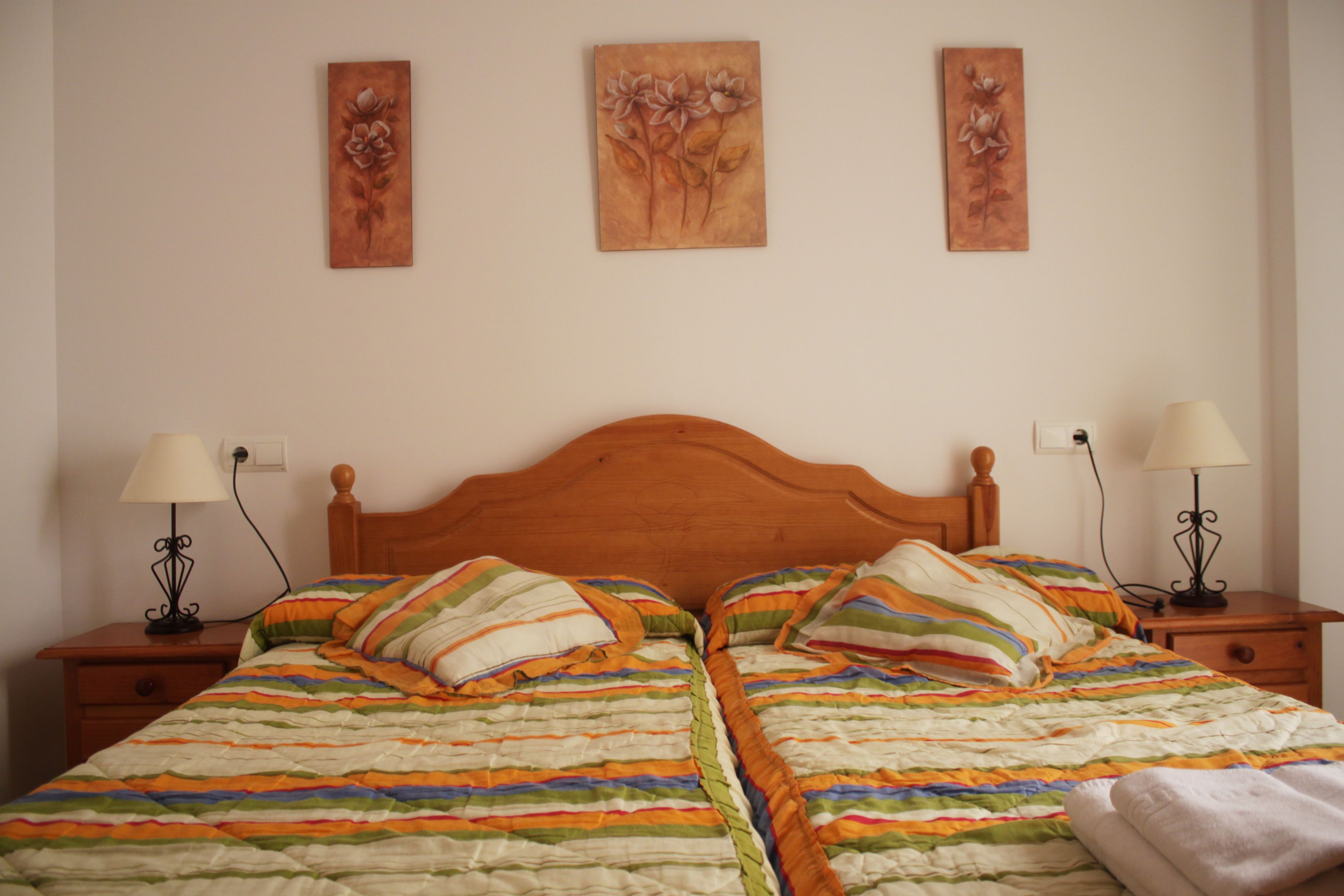 Un lit double avec une couvre-lit à rayures, deux tables de chevet avec des lampes et trois tableaux représentant des motifs floraux accrochés au mur.