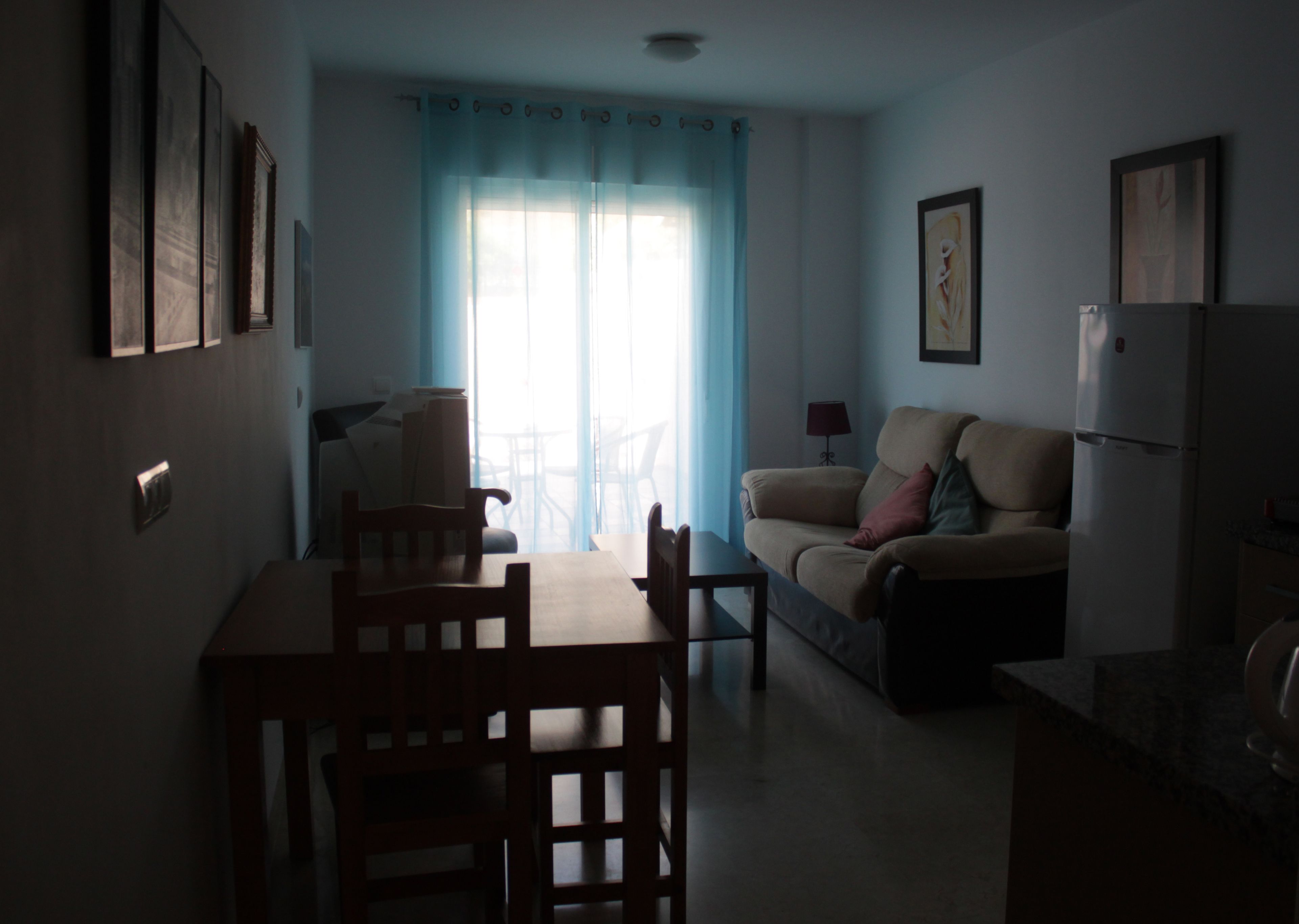 Una sala de estar con un sofá, una mesa de comedor con sillas, un refrigerador y una ventana con cortinas.