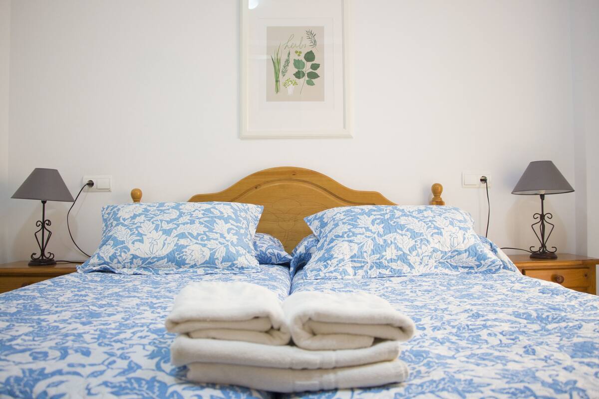 A neatly made bed with blue floral bedding, two bedside lamps, and a framed botanical artwork on the wall.