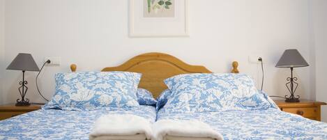 A neatly made bed with blue floral bedding, two bedside lamps, and a framed botanical artwork on the wall.