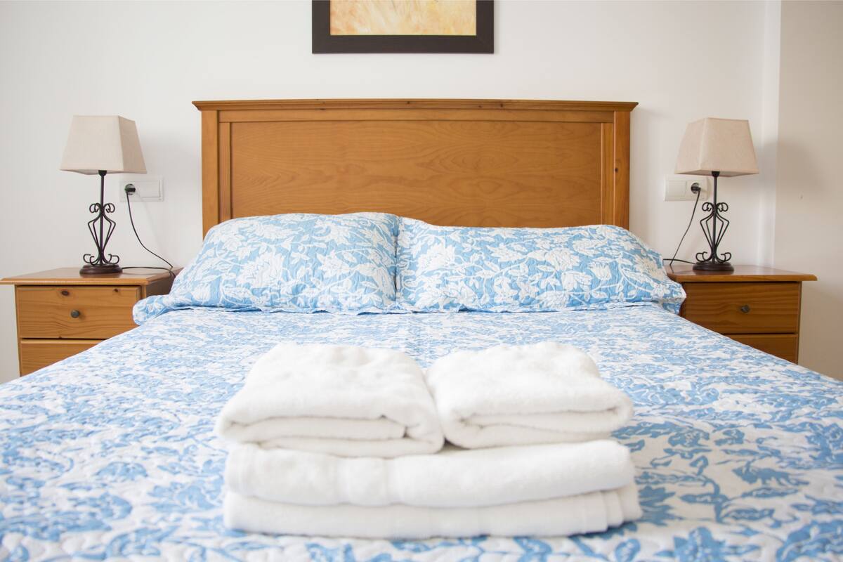 A neatly made bed with a floral-patterned bedspread and two pillows, flanked by bedside tables with lamps and a framed picture above the bed.