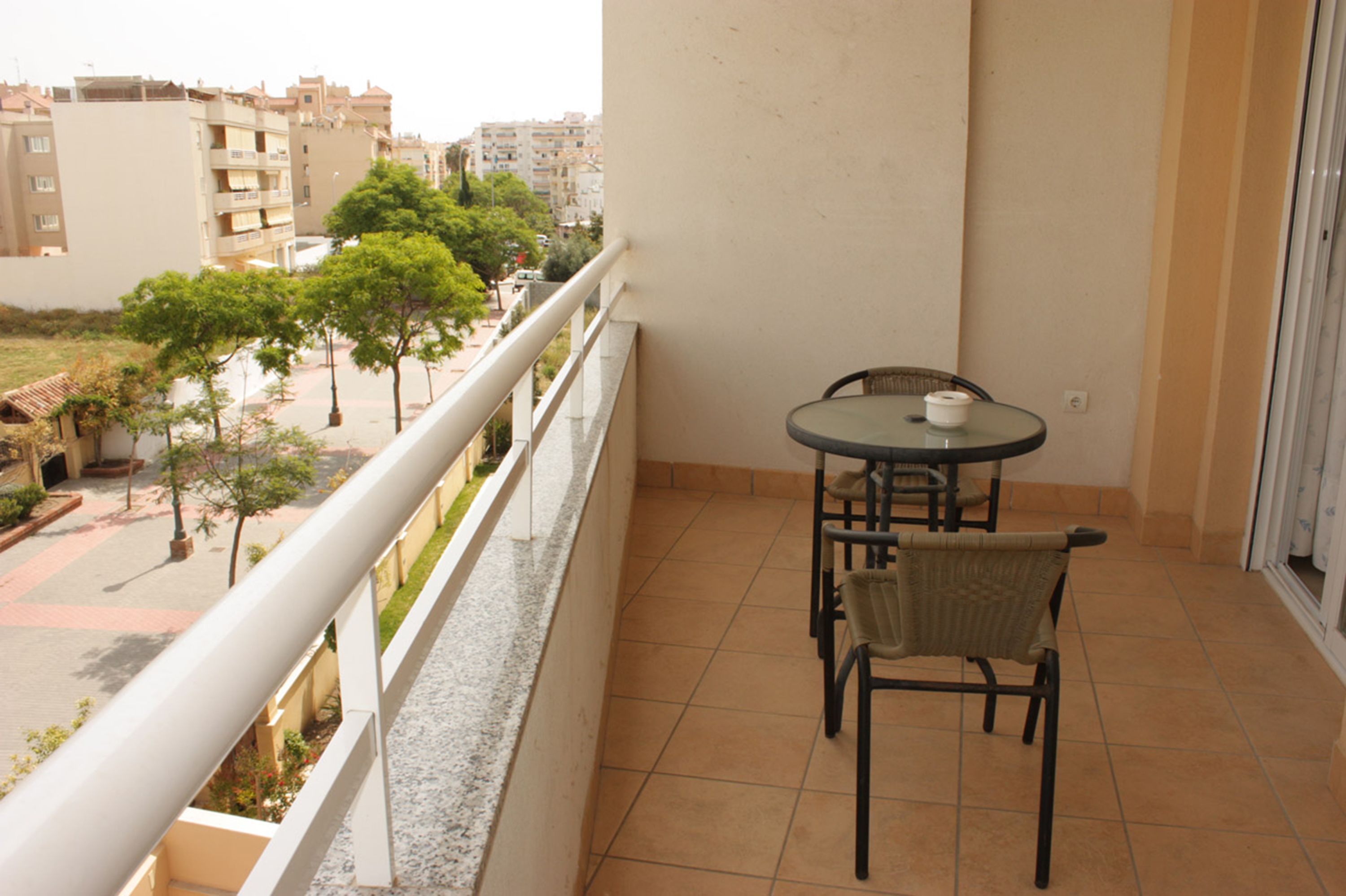 A balcony with a table and chairs, overlooking a street with trees and buildings.