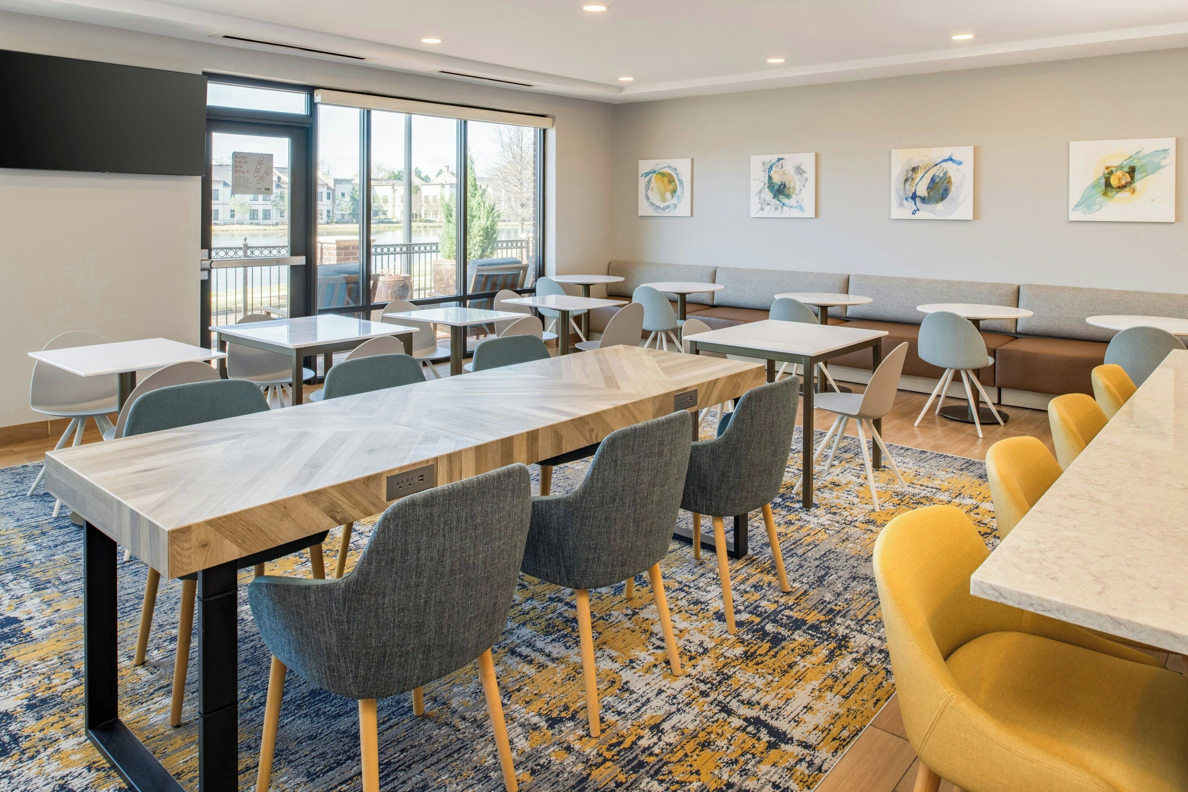 A modern dining area with multiple tables and chairs, large windows offering a view of the outdoors, and artwork on the walls.