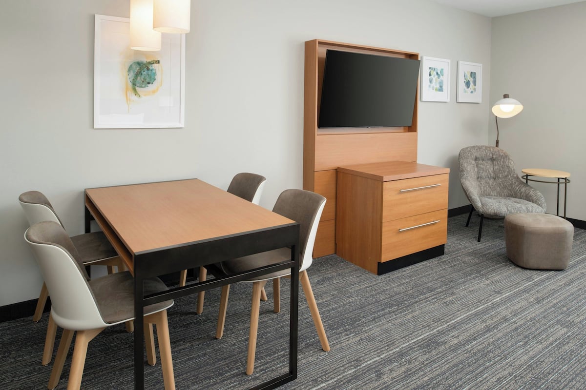 A modern hotel room with a wooden desk, a flat-screen TV, a grey armchair, and a small side table.