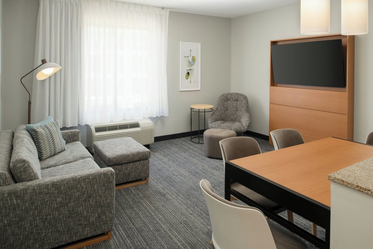 A modern hotel room with a grey sofa set, a wooden dining table, and a flat-screen TV mounted on the wall.
